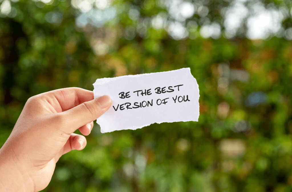 The image is of a hand, holding a note saying 'Be the best version of you'. The background is outside in nature with trees