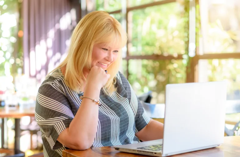 A woman smiling while working on a laptop in a bright café.