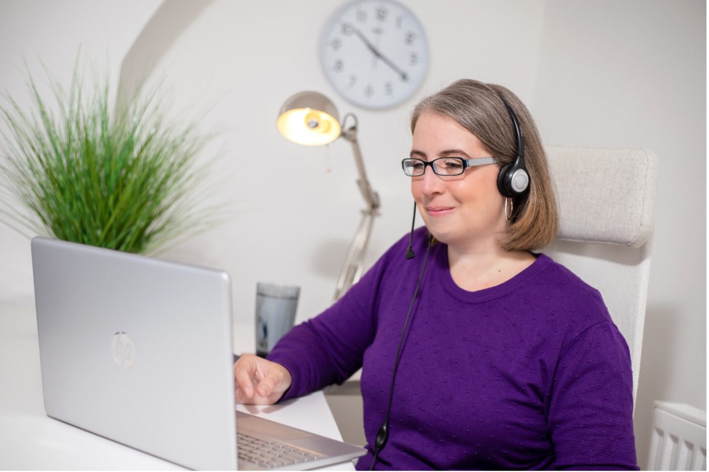 A photograph of me Hannah Paskin. I am in a purple top, with a headset on and looking at a laptop. It represents me in a coaching session.