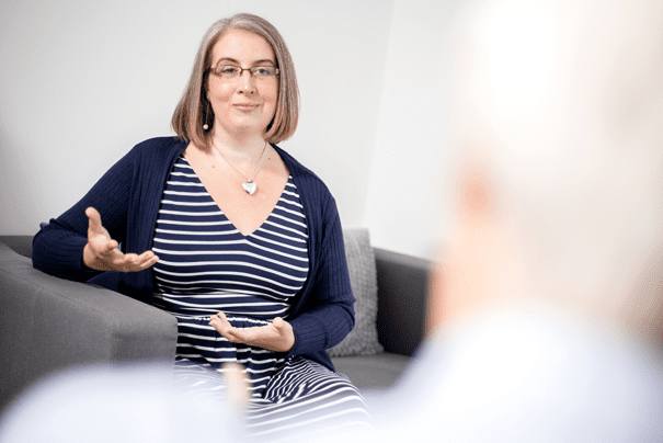 Image of me, Hannah Paskin, in a mock up Therapy session photoshoot. I am shown sat on a couch, I am wearing a navy and white stripped dress, and a navy cardigan. You can see the blurred back of a head of a mock client.