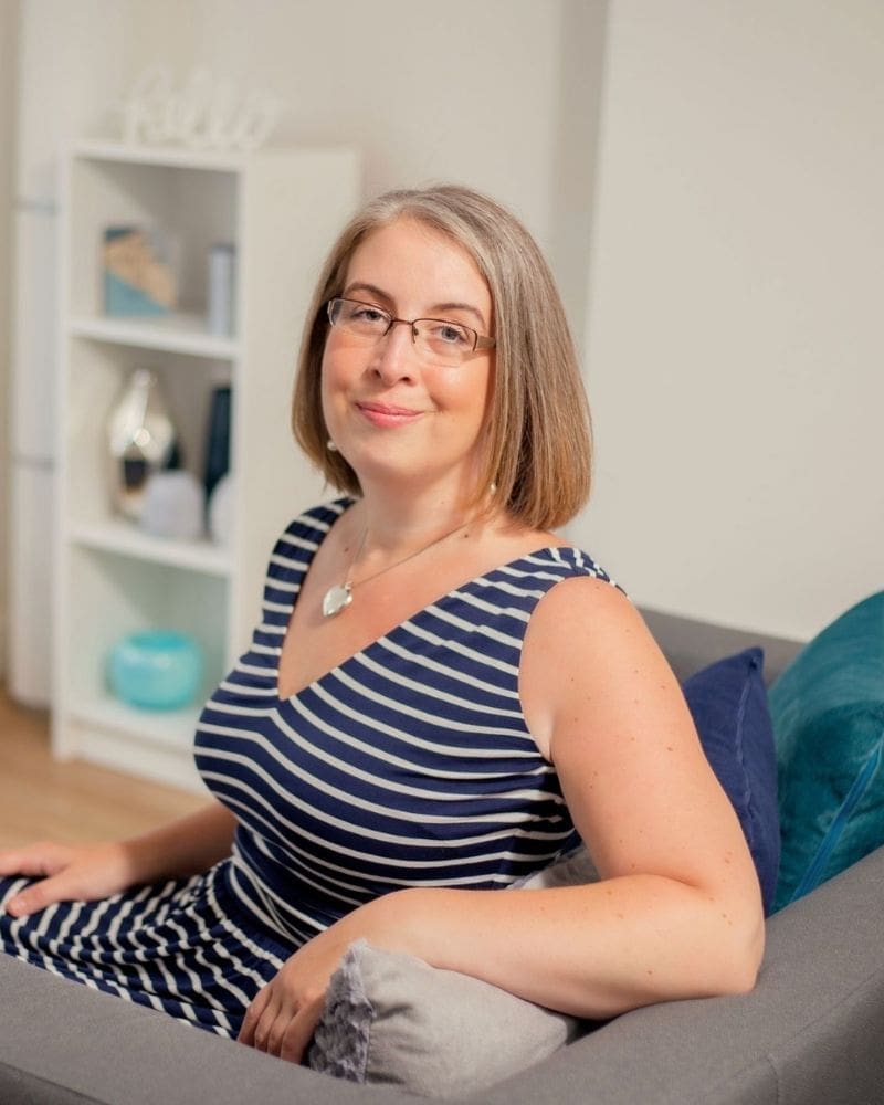 A photo of Hannah Paskin in her Therapy Clinic in Middlewich. She is sat on a sofa turning back to look at the camera. The sofa is grey with cushions on. Hannah is wearing a navy and white stripy dress and wears glasses.