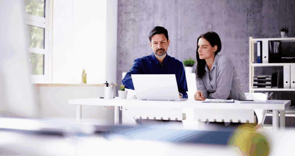 The photo contains and business man and woman sat at a table together looking at a laptop. He has dark hair a beard and a blue shirt. She has dark hair and wears a grey blouse. The desk is white and the background of the room is grey