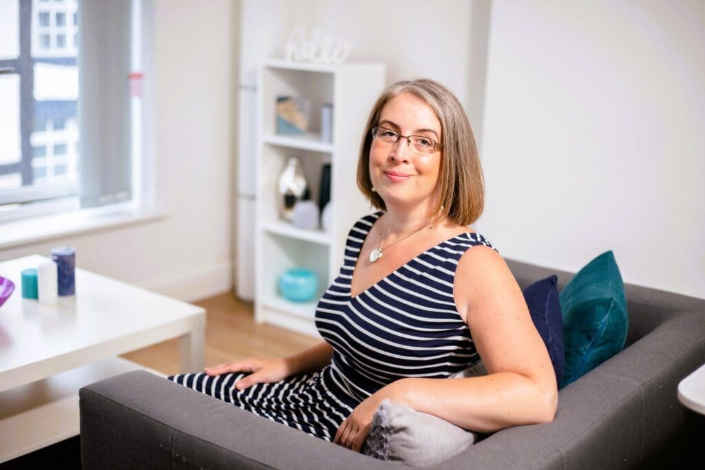 Hannah Paskin in her counselling room in Cheshire. She is posed for a photographer sat on a sofa. She is turned back towards the camera and smiling. In the background you can see her counselling room with a table and bookcase.