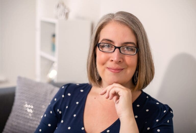 Hannah Paskin Psychotherapist. Head and shoulders shown. Her chin leans onto a curled hand. She is smiling at the camera. She wears a blue and white dress. Her therapy room can be seen out of focus in the background.