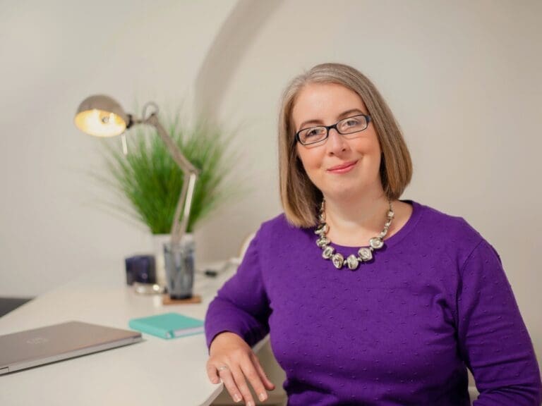 Photo shows therapist Hannah Paskin. She is smiling at the camera. She wears a purple jumper and a chunky silver necklace. She has a bob hair cut with mid brown colour hair. She wears blue glasses.