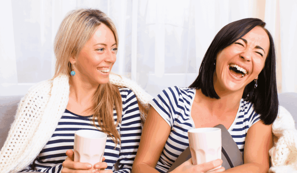 Two friends laughing and enjoying tea together on a sofa.