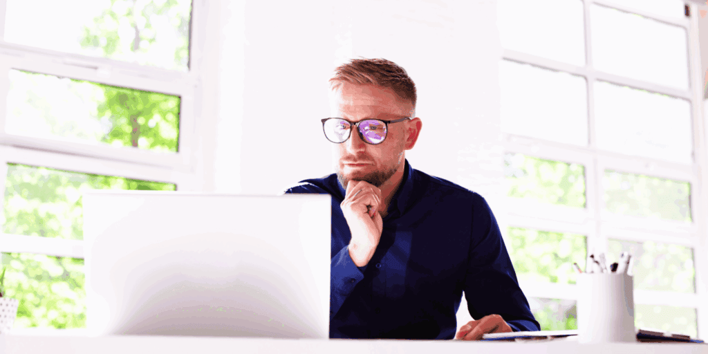 The photo shows a blonde haired male. He is sat at a desk with a laptop in front of him he is looking at. He wears glasses and has a nose ring, he wears a smart blue shirt. He has his head leaning onto a curled hand.