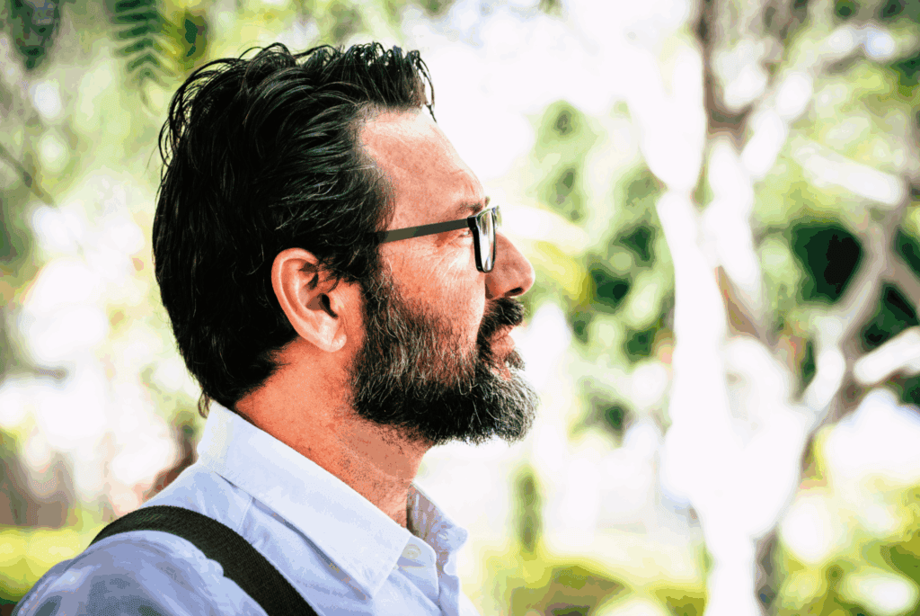 A man with a beard and glasses standing outdoors, surrounded by greenery.