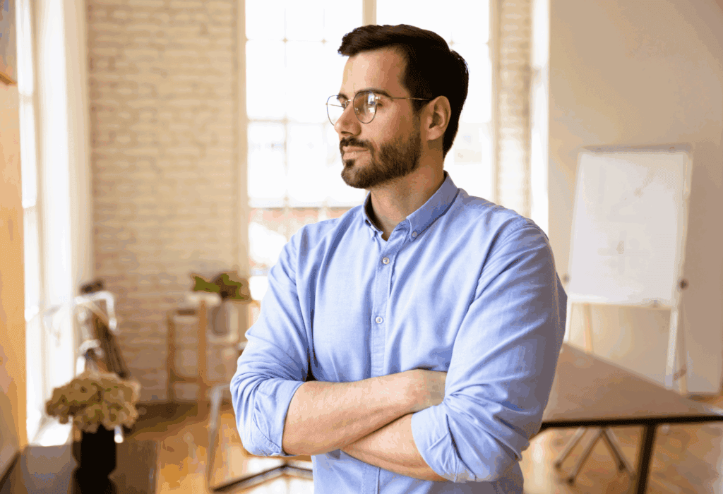 A man wearing glasses, looking contemplative in a bright office environment.