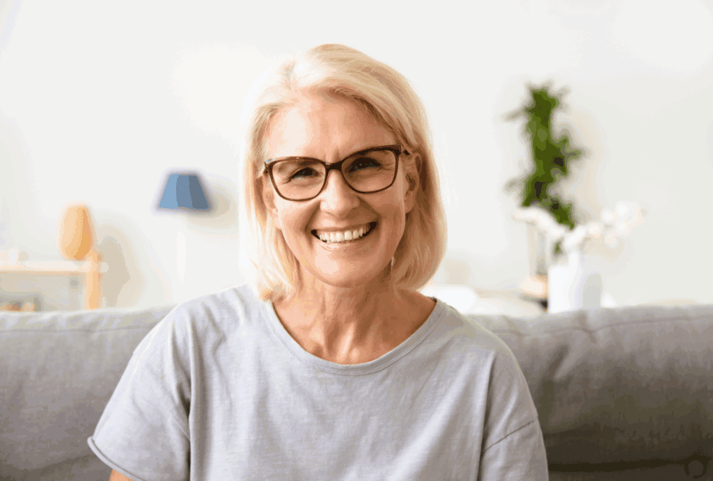 The photo shows a mature lady with white bob hair and red glasses frames. She is sat down and smiling at the camera. She is wearing a grey tshirt, and the sofa is also grey. The background is white