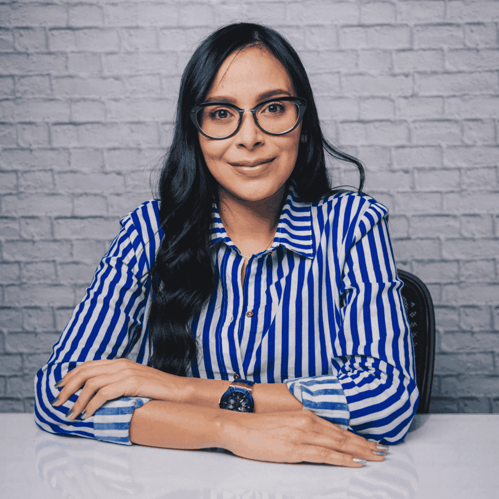 A professional woman in a blue and white striped shirt, seated at a desk with a brick wall background.