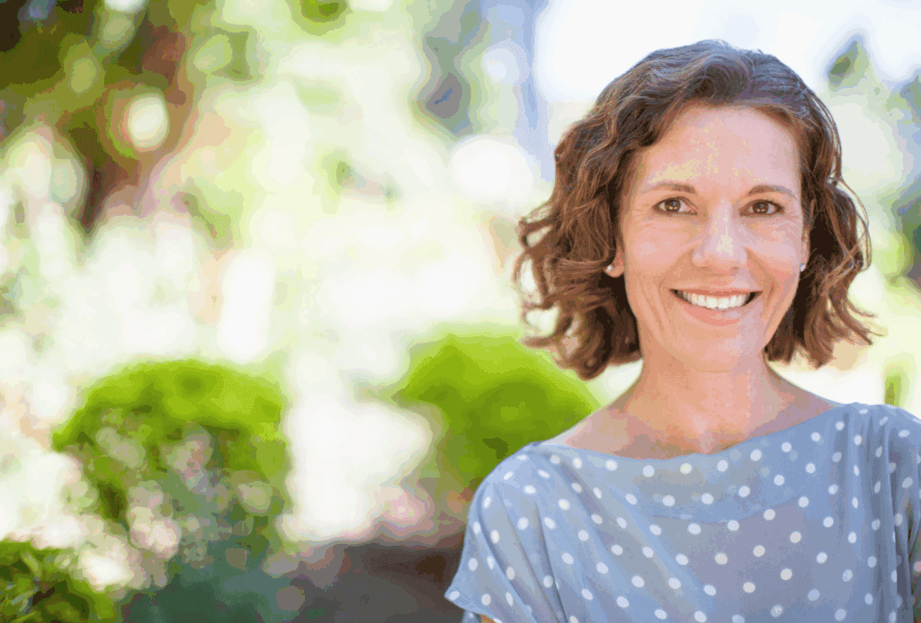 Middle aged lady posing for camera. She wears a pale blue top with white spots. She has bob length mid brown hair which his styled wavy. She smiles at the camera. The background is blurred out