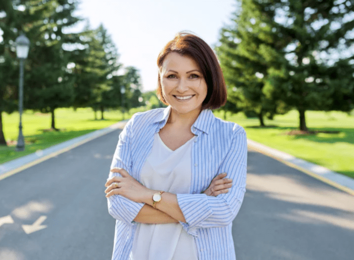 A cheerful woman smiling confidently in a park setting with trees in the background.