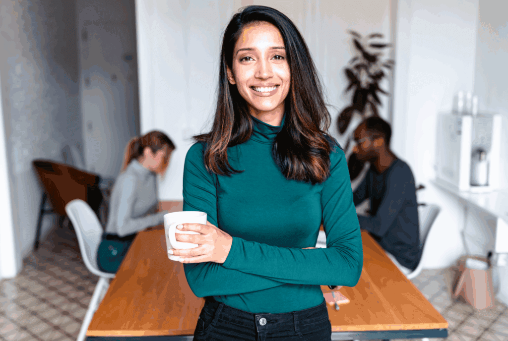 The photo shows a female in the foreground stood up. She has dark long hair, she has a turquoise top and black jeans. She is stood with arms folded and a cup in one hand. She smiles at the camera.