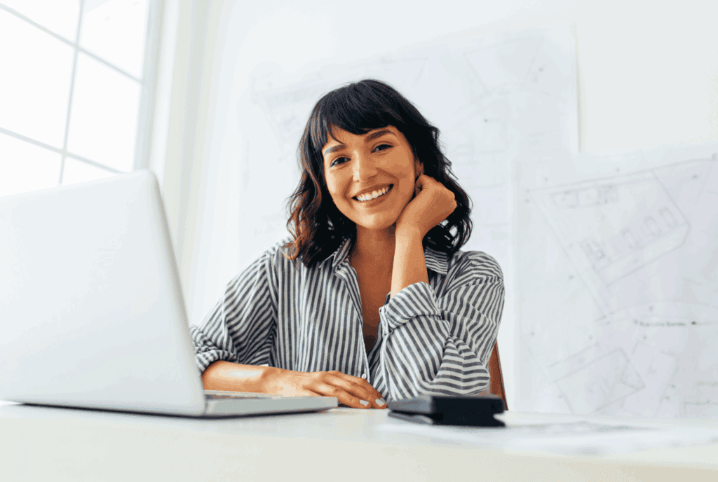 Female sat at a desk. The desk is white, with a white laptop. There also looks to be a calculator on the desk. She wears a stripy blouse and has dark wavy hair with fringe. She leans onto the desk with one arm