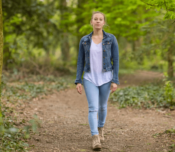 Female walking in forest. She is walking on a path. There are out of focus trees behind her. Her hair is blonde and tied up. She is wearing walking boots, jeans, a tshirt and denim jacket