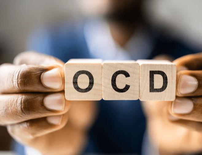 3 small wooden cubes are at the centre of the screen with the letters OCD on them. They are held between a persons fingers in a row. The person is out of focus in the background.