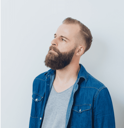 Male shown looking into the distance. He wears a grey tshirt with a denim shirt on top. He has a hair style mixed with shared sections and longer sections and has a groomed beard. The background is white.