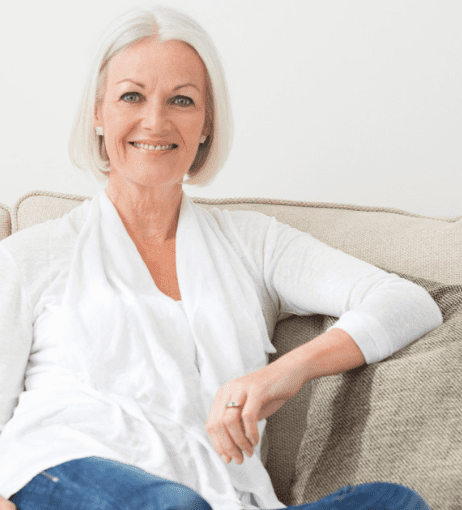 A mature lady sitting on a sofa. The sofa is cream and wall behind is white. She has white hair in a bob. She wears a white blouse with jeans. She has her arm resting on the armrest. She is smiling at the camera.