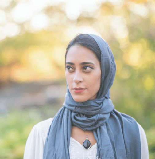A lady looking into the distance smiling. The background is out of focus but indicates lights and trees. The lady wears white with a grey head scalf.