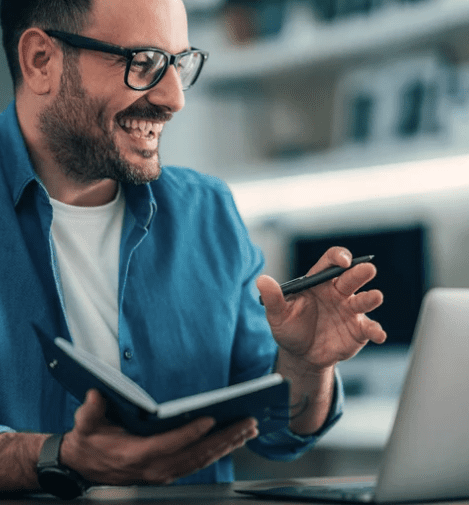 Male smiling and looking at laptop screen on desk in front of him. He wears a white tshirt and denim shirt. He has a watch on and glasses. In one hand he holds a notepad, and the other a pen.