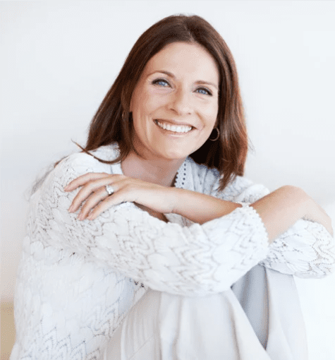 A female is sat on the floor with her knees to her chest. Her arms are loosely crossed. She is wearing a white top and white trousers. She has dark mid length hair. She is looking at the camera and smiling.