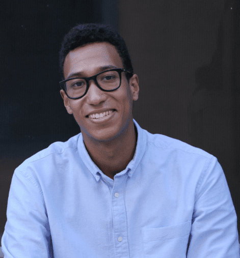 A man smiling at the camera. He has a blue shirt. He has dark hair and wears black glasses. There is a dark background with no other details visible. He is smiling at the camera.