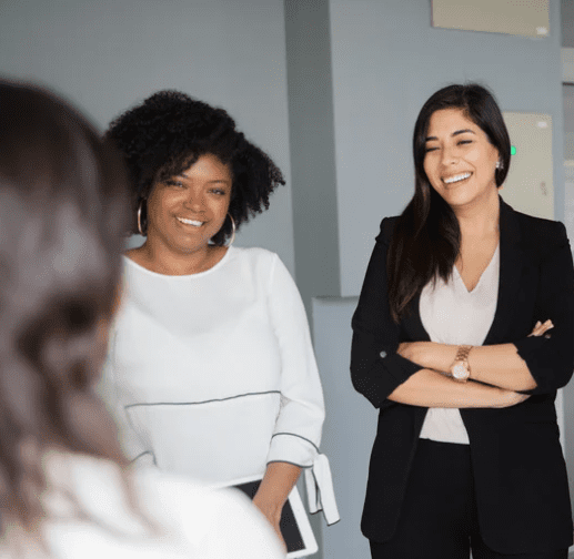 3 women stood chatting. One woman is shown only the back of her out of focus. 2 others are shown smiling at her. One holds an ipad. The other has her arms crossed. All 3 have dark hair.