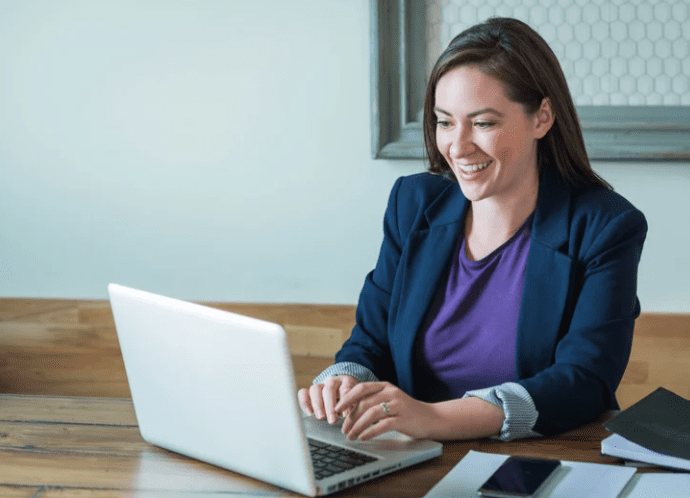 Female sat at wooden bench desk. She has dark hair and wears a purple top and blue jacket. On the desk is a grey laptop a phone and some paper. She is looking at her laptop with her hands hovering above it. She is smiling.