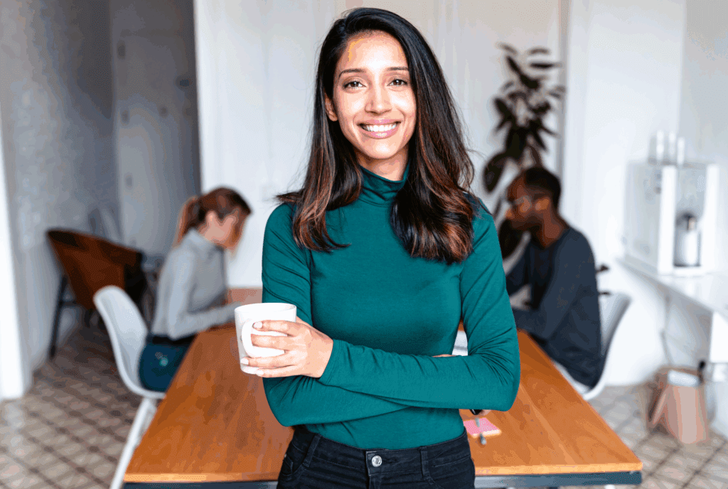 A stock image. The photo shows a female in the foreground stood up. She has dark long hair, she has a turquoise top and black jeans. She is stood with arms folded and a cup in one hand. She smiles at the camera.