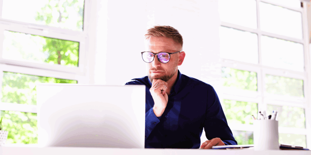 A stock image. The photo shows a blonde haired male. He is sat at a desk with a laptop in front of him he is looking at. He wears glasses and has a nose ring, he wears a smart blue shirt. He has his head leaning onto a curled hand.