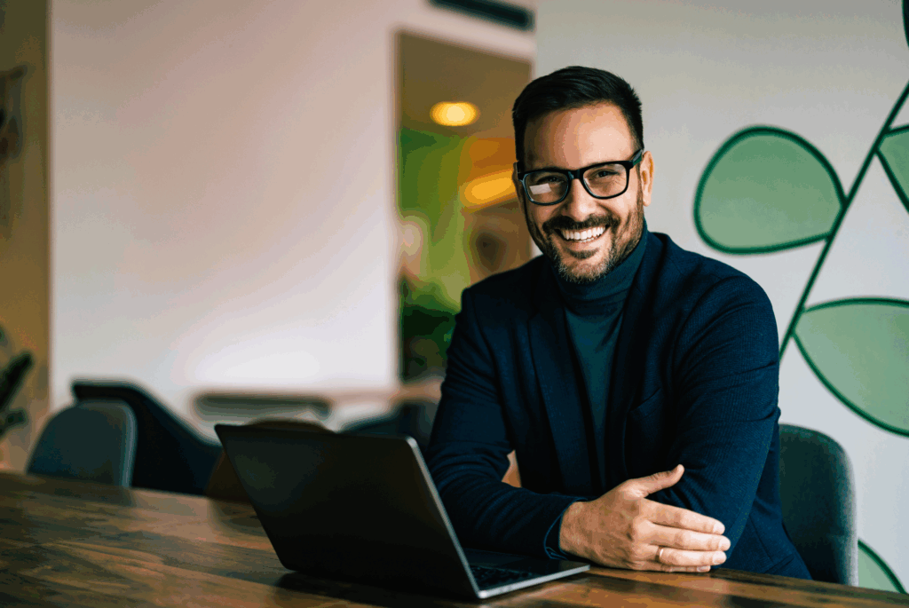 A stock image. A male smiling at the camera. He is sat at a desk with a laptop. He wears and roll necked top with a smart jacket and also wears glasses and has facial hair. In the background is an office, which has large patterns on the wall