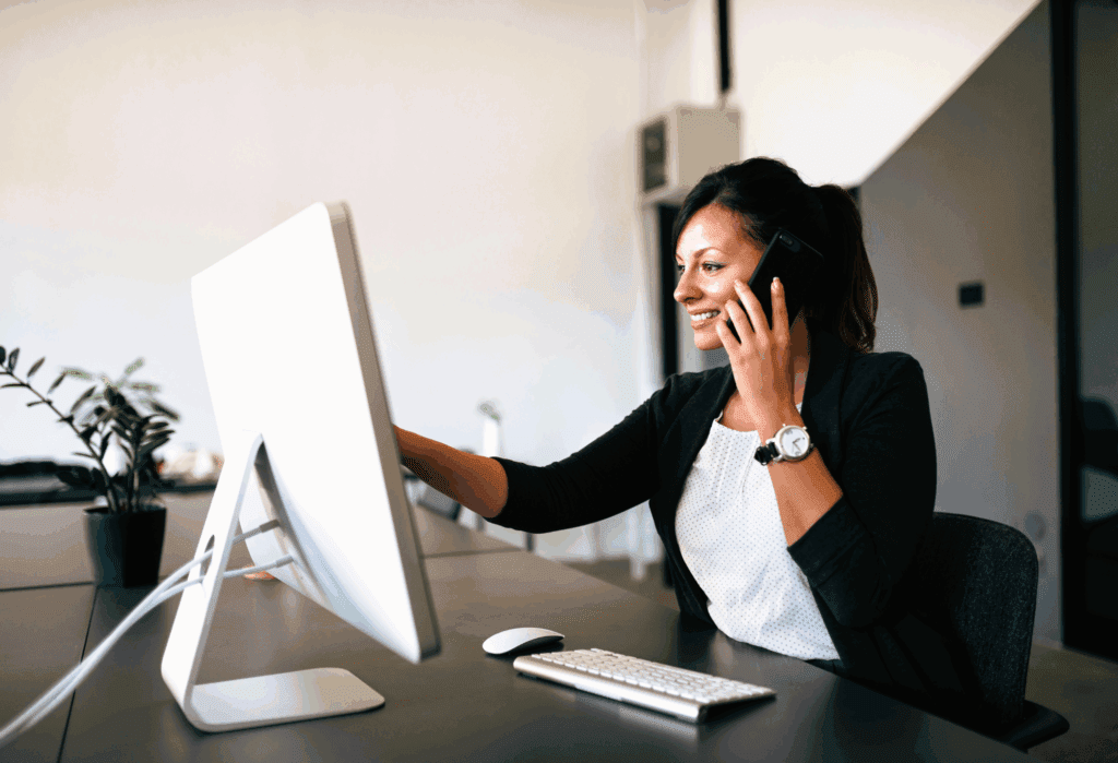 A female is sat at a desk in the office. The desk is long and dark. In front of her on the desk is a desktop computer screen, a keyboard and mouse. With one hand she holds her mobile phone to hear ear, with the other she points to the screen.