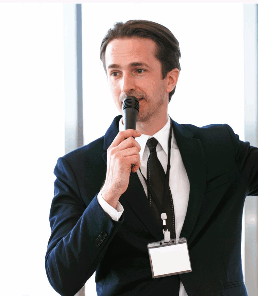 A male stood up. He is wearing a dark suit with white shirt and a dark tie. He appears to be in an office. He has a microphone in his hand he is talking to, and has a lanyard around his neck. It suggests he is speaking at an event.