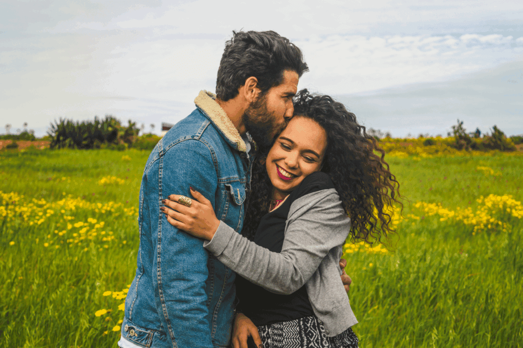 Image shows a couple that are male female. They are embracing. She is smiling, and he is kissing the top of her head. She faces the camera direction, he faces sides wards to her. They are stood in a field outside.
