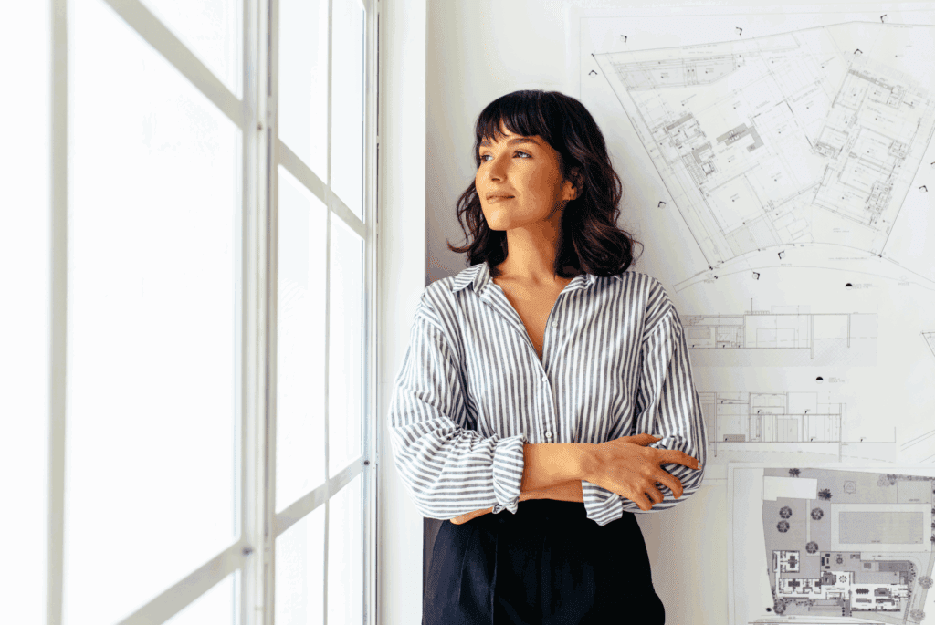 A woman is stood looking out of the window. The wall and window are white. She has dark hair that is curly with a fringe. She wears a stripy blouse and dark trousers. On the wall behind her seems to be drawn plans for buildings.