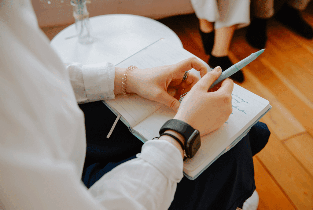 A zoomed in image of a female with a notepad on her lap. She has a hair bobble around her wrist. She holds a pen with one hind. A bud vase is on a side table nearby