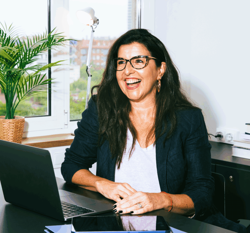 A stock photo. A lady at work laughing or smiling big. She is sat at a desk with a laptop. She is looking beyond the camera. She has dark thick long wavy hair and wears glasses. In the window ledge behind her is a plant and lamp