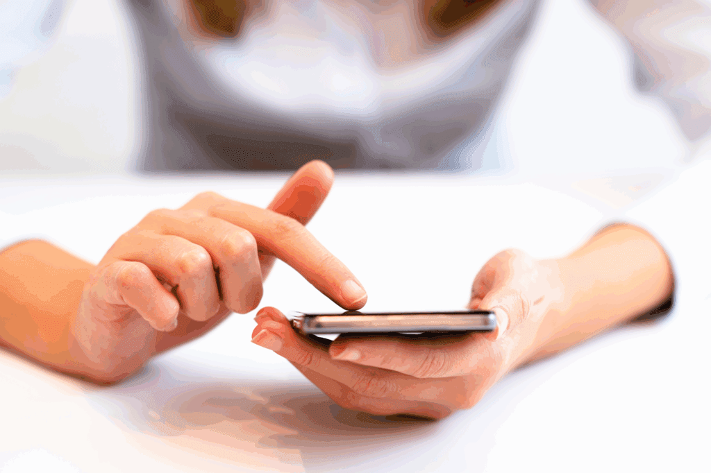 A zoomed in image of a female tapping her mobile phone. She is leaning on a white table as she does this. Her body is out of focus in the background but looks to be a white top and blonde hair