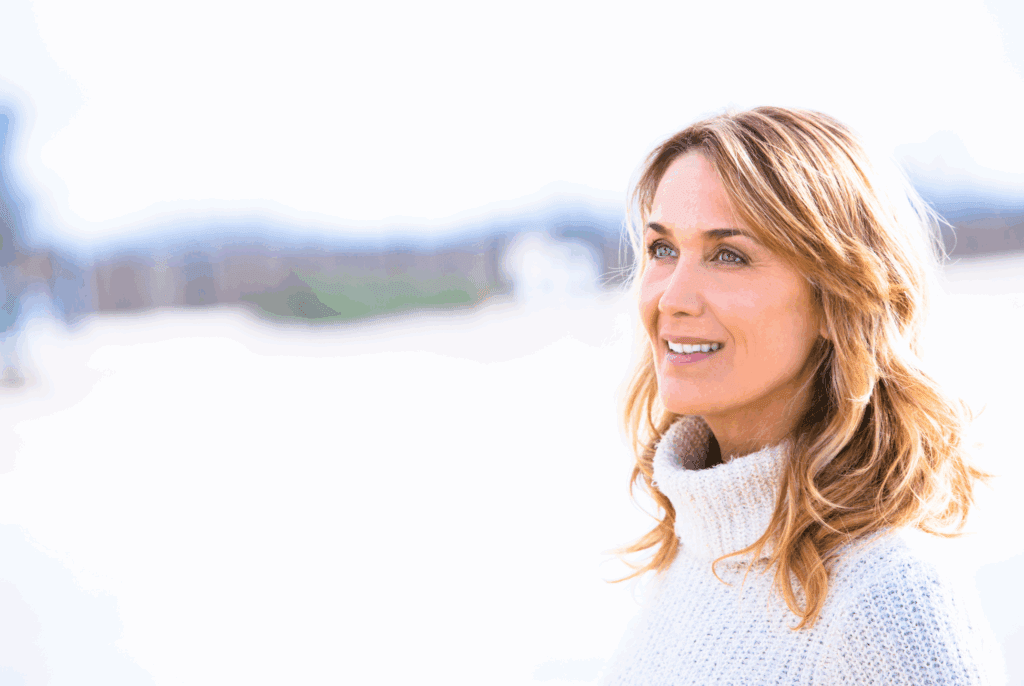 Female with white fluffy jumper with roll neck. She has blond hair with loose curls. She is smiling and looking into the distance. The background is out of focus.