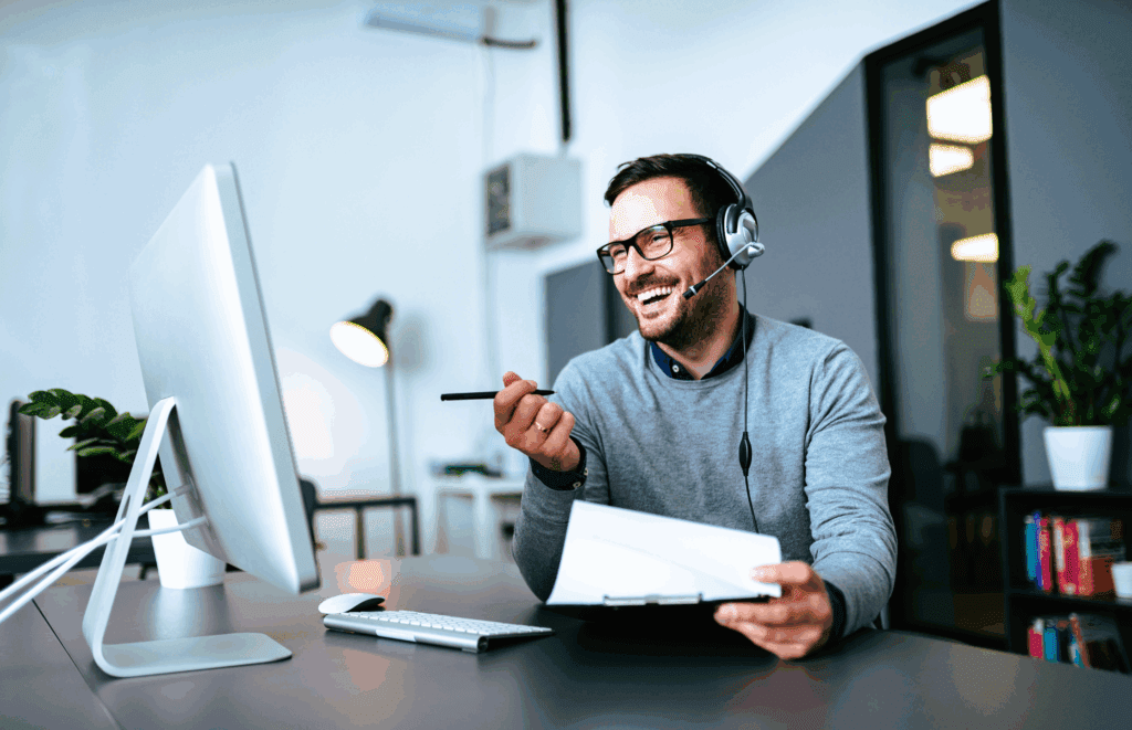 Male in office. He is sat at a desk. He is looking at a laptop off the image. In one hand he has a pen, in the other a clipboard. He also wears a headset. He wears grey and is smiling