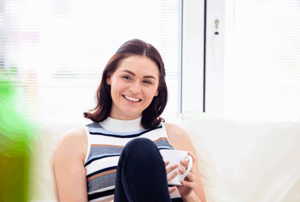 Female sat on sofa with a cup of hot drink. Behind her is a window. She is sat with one leg up on the sofa. She wears a sleeveless stripy top and blue leggings. She has dark hair part tied up. She is looking at someone not in the image