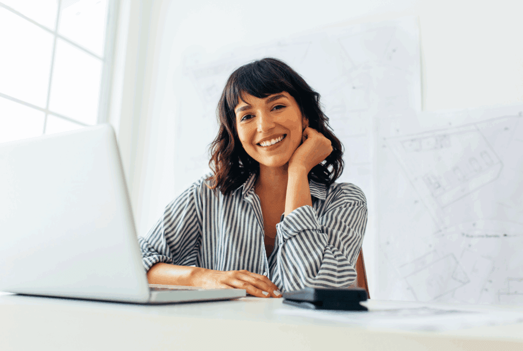 Female sat at a desk. The desk is white, with a white laptop. There also looks to be a calculator on the desk. She wears a stripy blouse and has dark wavy hair with fringe. She leans onto the desk with one arm