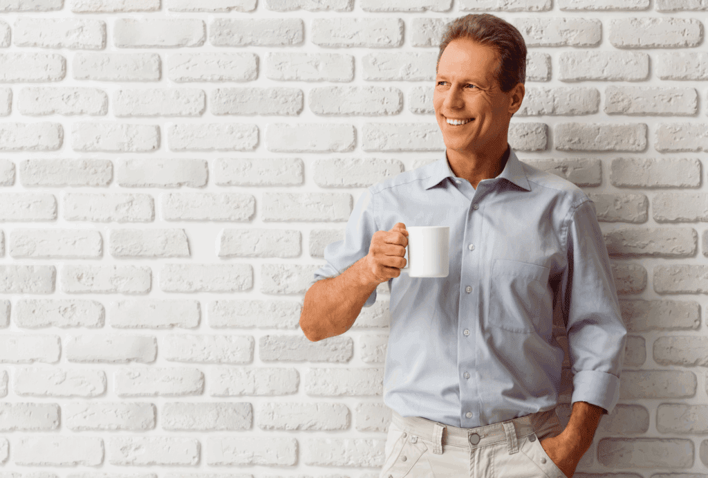 Male stood against brick wall painted white. He has mid brown hair and is smiling a big smile. He has a pale blue shirt on and white trousers. He holds a white mug in his hand and looks into the distance.