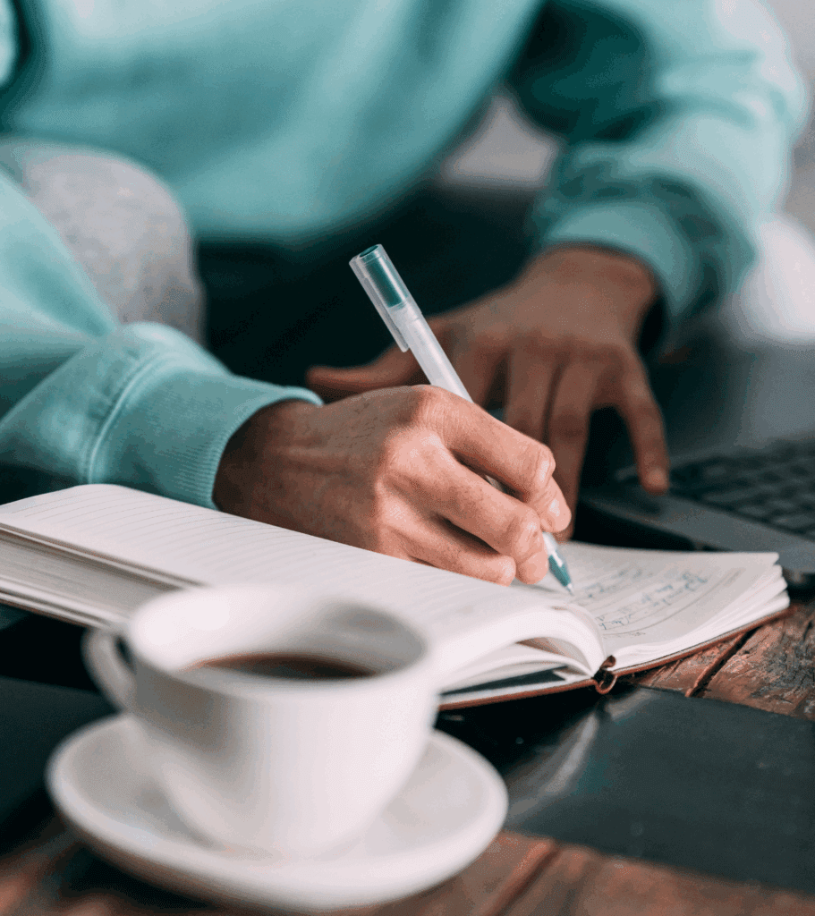 A close up of someone writing in a notepad. They are sat on a sofa leaning forwards and their notepad is on a low table. There is also a mug on the table. The person holds a pen and writes with 1 hand, the other hand on their lap.