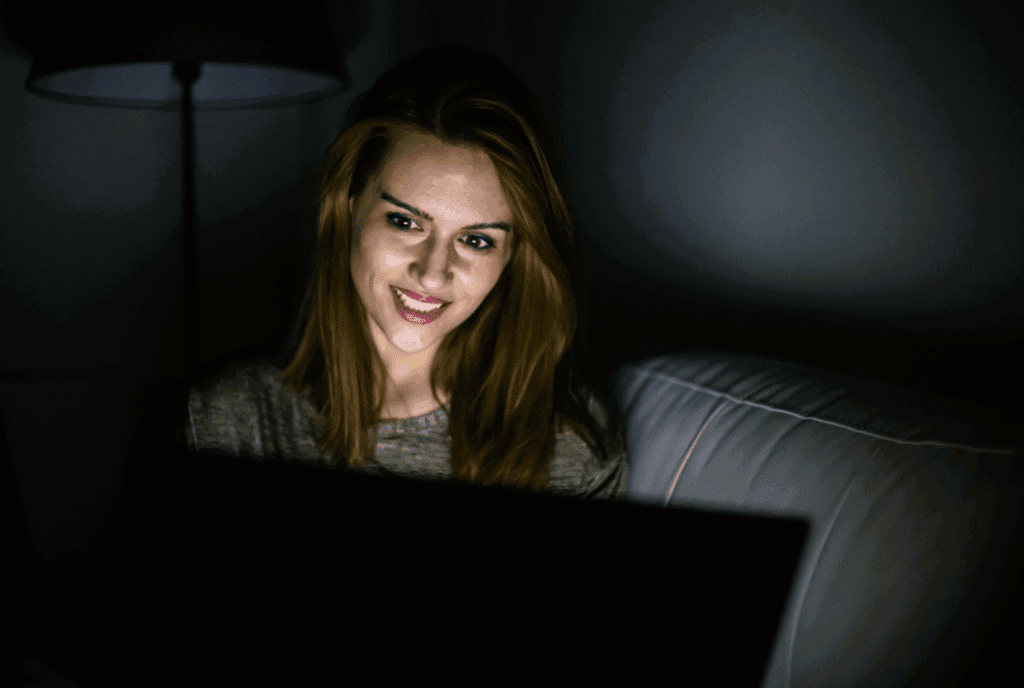 Female sat on sofa working on laptop in the dark. Her face is illuminated by laptop screen. She has auburn hair, a grey top and wears makeup. The background is in darkness.