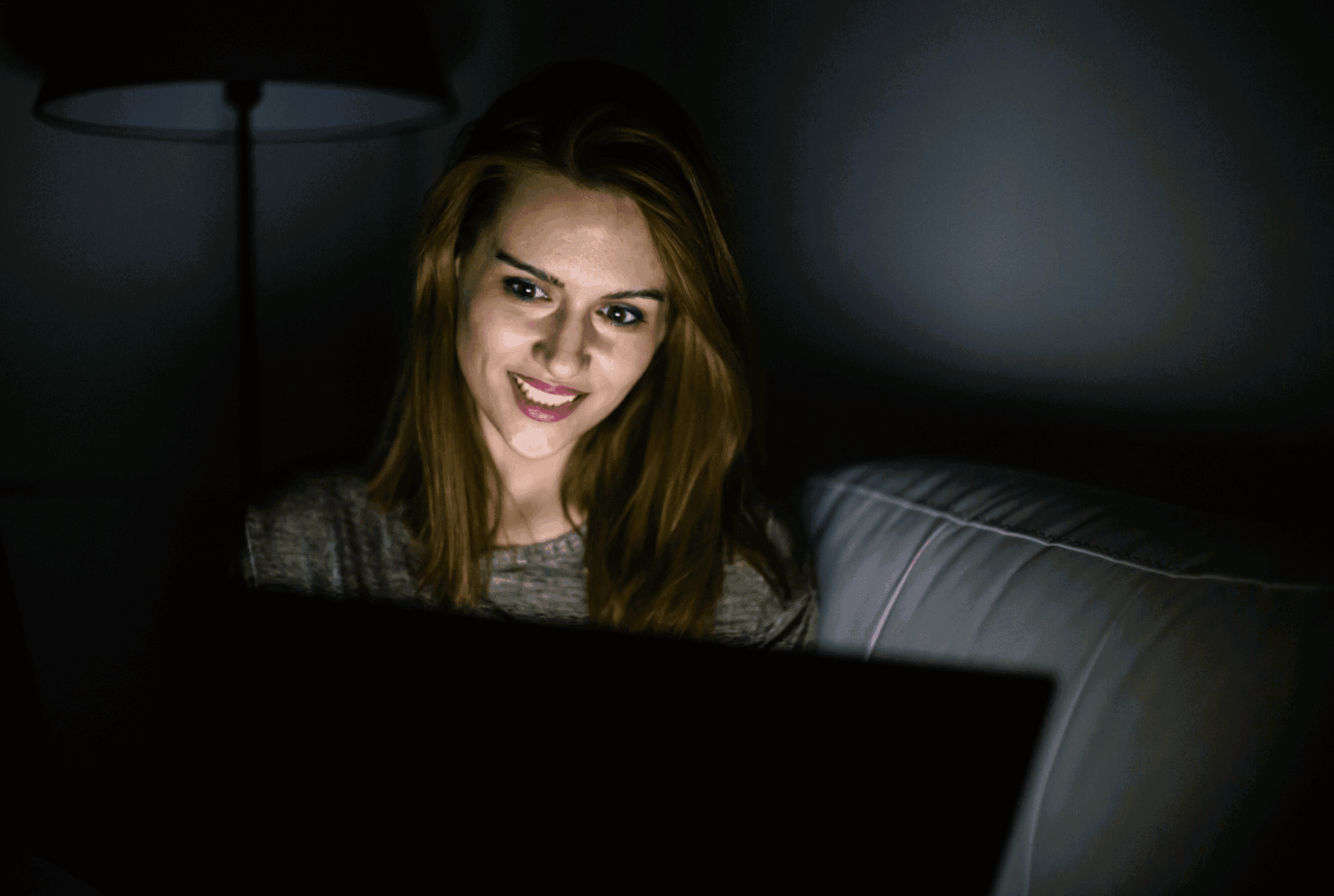 Female sat on sofa working on laptop in the dark. Her face is illuminated by laptop screen. She has auburn hair, a grey top and wears makeup. The background is in darkness.