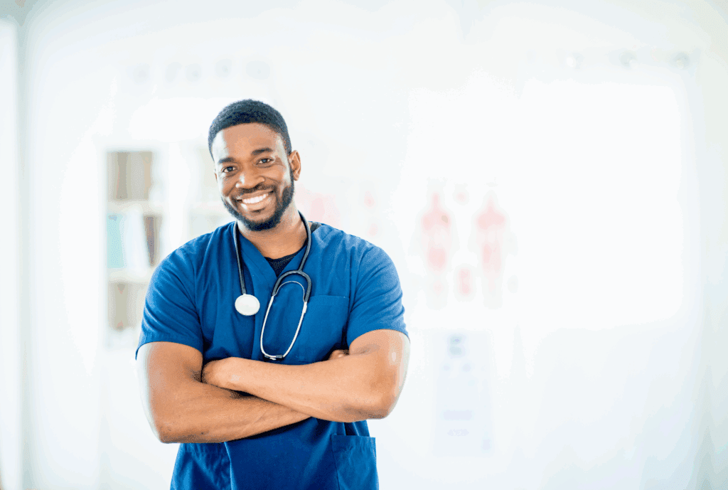Image of male doctor in scrubs. His uniform is blue. He has his arms crossed. He has a medical device around his neck. He is looking at the camera and smiling
