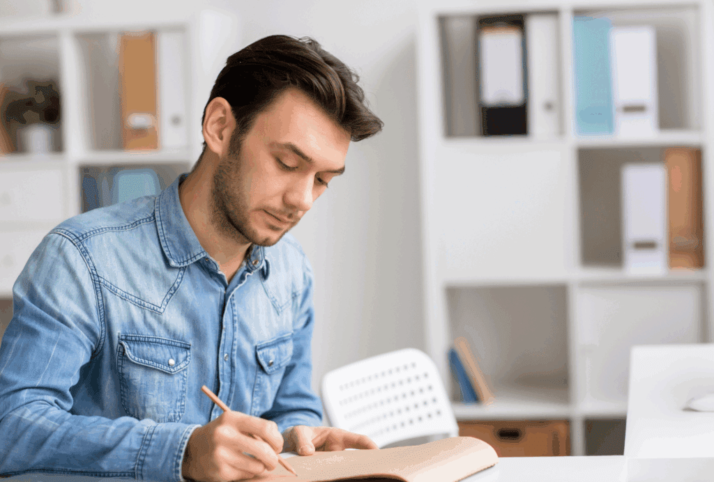 A male sat at a desk. He is writing in a notebook on the desk. He wears denim and has dark hair.