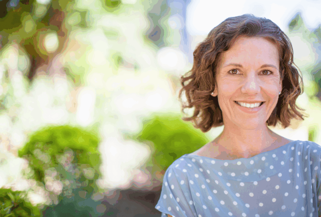 Middle aged lady posing for camera. She wears a pale blue top with white spots. She has bob length mid brown hair which his styled wavy. She smiles at the camera. The background is blurred out